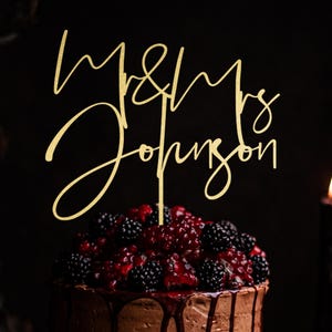 May include: A gold-colored cake topper with the words "Mr & Mrs Johnson" in a cursive font. The topper is placed on a chocolate cake decorated with blackberries and a red sauce. The cake is on a dark background.