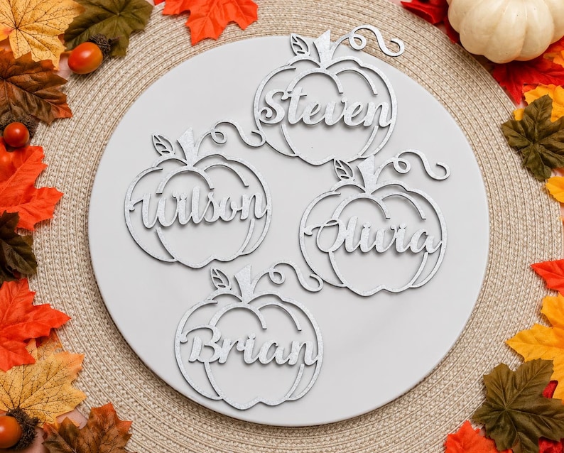 Puede incluir: Cuatro adornos blancos con forma de calabaza cortados con l&aacute;ser, cada uno con un nombre diferente en letra cursiva. Los nombres son Steven, Wilson, Olivia y Brian. Cada adorno de calabaza incluye un tallo y un detalle de zarcillo.