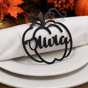 Puede incluir: Un servilletero negro mate con forma de calabaza que presenta el nombre Olivia en letra cursiva. El dise&ntilde;o recortado incluye un tallo y una hoja en la parte superior, apoyado sobre una servilleta de tela blanca en un plato blanco.