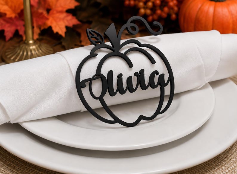 Puede incluir: Un servilletero negro mate con forma de calabaza que presenta el nombre Olivia en letra cursiva. El dise&ntilde;o recortado incluye un tallo y una hoja en la parte superior, apoyado sobre una servilleta de tela blanca en un plato blanco.