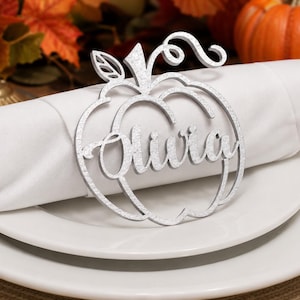 Puede incluir: Un servilletero plateado con purpurina en forma de calabaza que presenta el nombre Olivia en letra cursiva. El dise&ntilde;o recortado incluye un tallo y una hoja en la parte superior, rodeando la servilleta de tela blanca sobre un plato.
