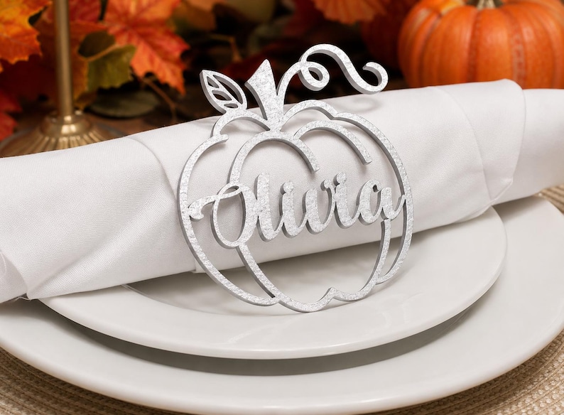Puede incluir: Un servilletero plateado con purpurina en forma de calabaza que presenta el nombre Olivia en letra cursiva. El dise&ntilde;o recortado incluye un tallo y una hoja en la parte superior, rodeando la servilleta de tela blanca sobre un plato.