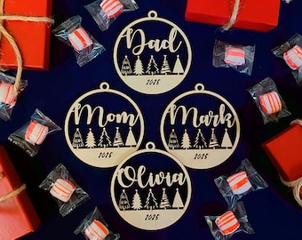 Puede incluir: Adornos navideños de madera con los nombres "Dad", "Mom", "Mark" y "Olivia" y el año 2025. Los adornos están rodeados de cajas de regalo rojas atadas con hilo y caramelos envueltos individualmente sobre una superficie azul oscuro.