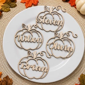 Puede incluir: Cuatro adornos de madera cortados con l&aacute;ser en forma de calabaza, cada uno con un nombre &uacute;nico en letra cursiva: Steven, Wilson, Olivia y Brian. Estos recortes de madera clara tienen forma de calabaza con tallos y zarcillos.
