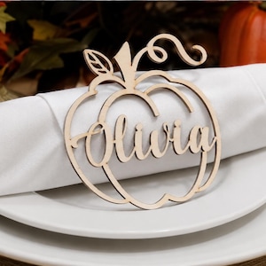 May include: A wooden pumpkin-shaped place card holder, laser-cut with the name "Olivia". The light brown place card sits on a white napkin and plate. The background is blurred, with autumn colours and pumpkins.
