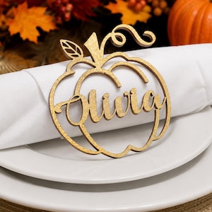 Puede incluir: Un servilletero de madera de color marr&oacute;n claro cortado con l&aacute;ser en forma de calabaza con el nombre Olivia en letra cursiva. El dise&ntilde;o de la calabaza incluye una peque&ntilde;a hoja y un tallo rizado, sobre una servilleta blanca.