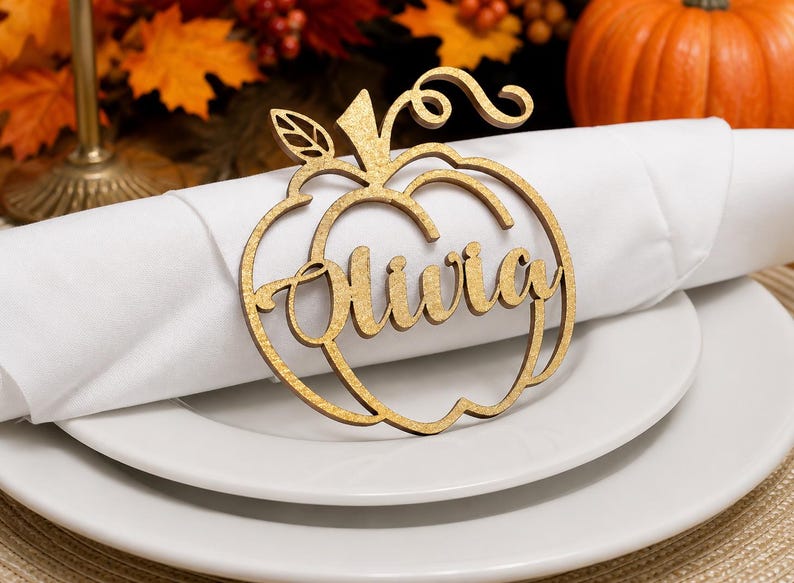 Puede incluir: Un servilletero de madera de color marr&oacute;n claro cortado con l&aacute;ser en forma de calabaza con el nombre Olivia en letra cursiva. El dise&ntilde;o de la calabaza incluye una peque&ntilde;a hoja y un tallo rizado, sobre una servilleta blanca.