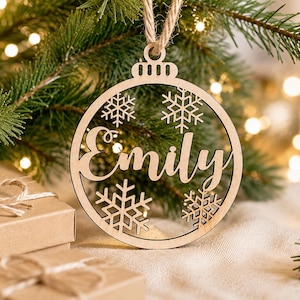 May include: A circular light-coloured wooden ornament featuring the name Emily cut out in the centre, surrounded by four intricate snowflake designs. The ornament is suspended by a piece of twine and has a small loop at the top for hanging.