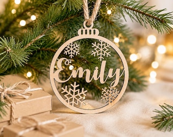 Puede incluir: Un adorno circular de madera de color claro con el nombre Emily recortado en el centro, rodeado por cuatro intrincados diseños de copos de nieve. El adorno está suspendido por un cordel y tiene un pequeño lazo en la parte superior.