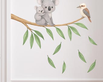 May include: Watercolor wall decal featuring a mother koala and baby koala sitting on a branch with a kookaburra. Green leaves are falling from the branch. The nursery decor also includes a white crib and dresser.