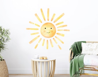 May include: A watercolor-style wall decal of a smiling sun with yellow and orange rays. The sun is on a white wall, with a small wooden table and a green throw blanket on a wicker chair in the background.