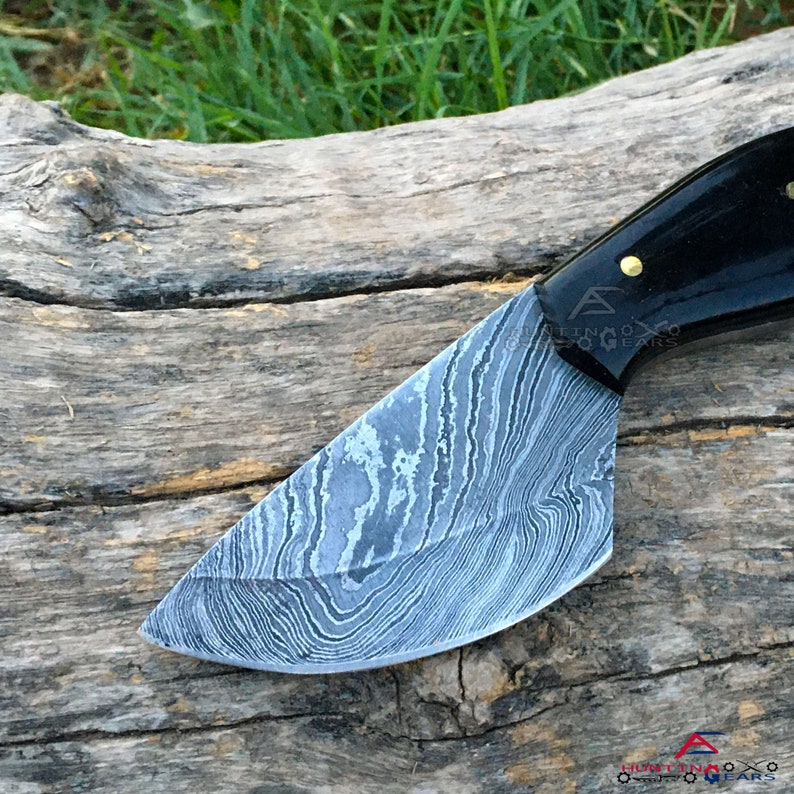 May include: A close-up of a Damascus steel knife with a black handle. The blade has a wavy pattern and the handle has a gold accent.