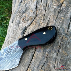 May include: A black knife with a Damascus steel blade and a black handle with gold accents. The handle has a hole for a lanyard.