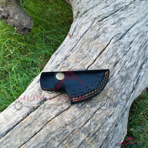 May include: A black leather sheath with a decorative stitching pattern and a brass button. The sheath is lying on a piece of weathered wood.