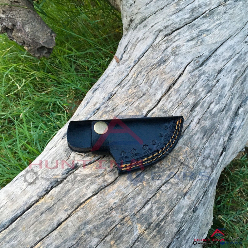 May include: A black leather sheath with a decorative stitching pattern and a brass button. The sheath is lying on a piece of weathered wood.