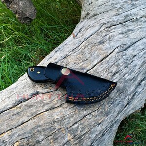 May include: A black leather knife sheath with a decorative stitching pattern. The sheath is lying on a piece of weathered wood.