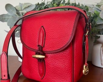 May include: A red leather crossbody bag with a flap closure. The bag features a textured surface, a braided accent, and a gold-tone clasp. The adjustable strap and trim are a darker shade of red. A small gold charm hangs from the side.