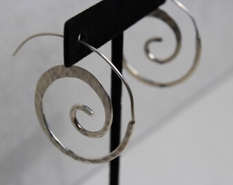 May include: Silver spiral earrings with a hammered texture. The earrings are displayed on a black stand against a white background. The design features a large, open spiral shape, creating a unique and eye-catching accessory.