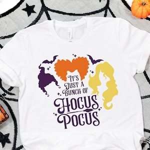 May include: White t-shirt with the phrase "It's Just a Bunch of Hocus Pocus" and silhouettes of three witches' hairstyles in purple, orange, and yellow. Halloween-themed with pumpkins and spiderweb decor.