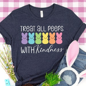Może przedstawiać: Koszulka w kolorze granatowym z napisem "Treat all peeps with kindness" i rzędem tęczowych pianek marshmallow Peeps. Koszulka leży na r&oacute;żowo-białym obrusie w kratkę z parą uszu kr&oacute;liczych, gniazdem z jajkami wielkanocnymi i wiązką zielonej trawy.