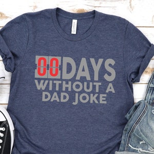 May include: A navy blue t-shirt with the text "00 DAYS WITHOUT A DAD JOKE" in gray and red. The shirt is paired with blue jeans, a black watch, and black canvas sneakers with white laces.