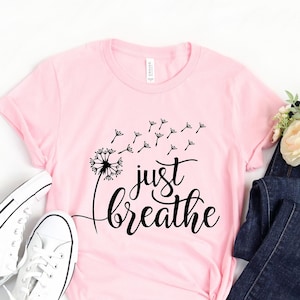 Op de afbeelding: Een roze t-shirt met een zwarte paardenbloem afbeelding en de tekst "just breathe" in een cursief schrift.