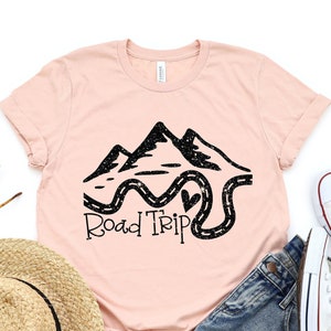 Puede incluir: Una camiseta rosa con un gr&aacute;fico negro de monta&ntilde;as y un camino sinuoso con el texto "Road Trip".