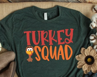 May include: A green t-shirt with the text "TURKEY SQUAD" in orange and red letters. A cartoon turkey with a red wattle is to the left of the text. The shirt is lying on a brown wooden surface with a pair of brown boots, a white cotton boll, and a brown flower.
