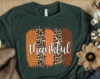 May include: A green t-shirt with a graphic design of orange and brown glitter stripes with a leopard print design and the word "thankful" in white lettering.