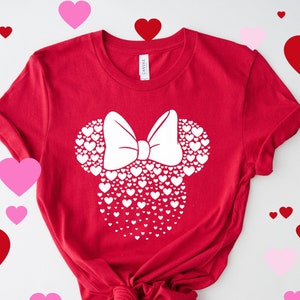 May include: A red short-sleeved t-shirt featuring a white graphic of a mouse head silhouette composed of numerous small hearts, topped with a large white bow. The shirt has a crew neckline and is displayed with a knotted hem at the bottom.