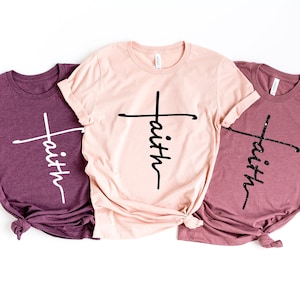 May include: Three t-shirts with the word "faith" written vertically with a cross in the middle. The shirts are in three different colors: maroon, pink, and light maroon.
