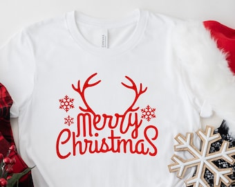 Puede incluir: Camiseta blanca con un gráfico rojo de "Feliz Navidad" con cuernos de reno y copos de nieve.