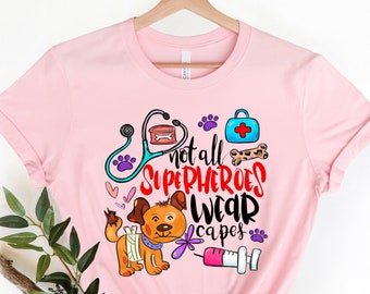 May include: Pink t-shirt featuring a cartoon dog wearing a bandage and a stethoscope. The text on the shirt reads "Not all superheroes wear capes".