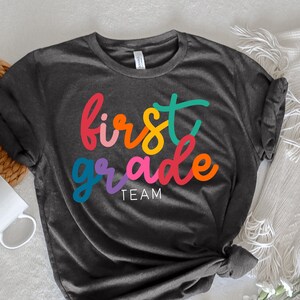 K&ouml;nnte beinhalten: Ein dunkelgraues T-Shirt mit den Worten "first grade team" in Regenbogenfarben.