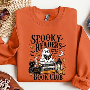 May include: An orange crewneck sweatshirt featuring a graphic print with the text SPOOKY READERS BOOK CLUB. The design includes a ghost wearing glasses reading a book atop a stack of books, a black cat, a mug, a lantern, and a pumpkin.