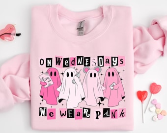 May include: A light pink crewneck sweatshirt featuring a graphic of four ghosts. The text reads "ON WEDNESDAYS WE WEAR PINK". One ghost holds a "BOO BOOK", another a phone, one has mouse ears, and the fourth holds an envelope. Heart-shaped glasses and lollipops are nearby.