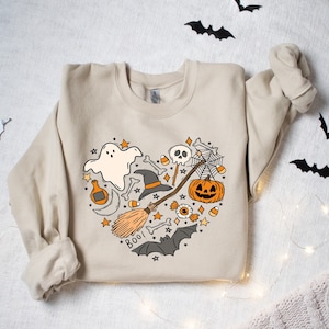 Może przedstawiać: Beżowa bluza z sercowatym wzorem przedstawiającym r&oacute;żne symbole Halloween, w tym ducha, kapelusz czarownicy, miotłę, dynię, czaszkę i napis "BOO!"