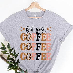 May include: Gray t-shirt with the text "But first, coffee" printed in black and brown. The word "coffee" is repeated three times, with each word featuring a different color and a leopard print design.