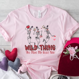 May include: Pink t-shirt with a graphic of three skeletons dancing and holding hearts. The text "Wild Thing You Make My Heart Sing" is printed below the skeletons.