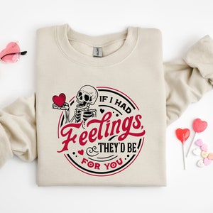 May include: A beige sweatshirt featuring a graphic print of a skeleton holding a heart. The graphic reads "If I had feelings, they'd be for you".