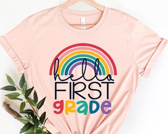 May include: A pink t-shirt with a rainbow and the text "hello first grade" in black and colorful letters.