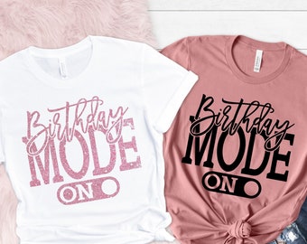 May include: Two t-shirts with the text "Birthday Mode On" in pink glitter and black lettering. The shirt on the left is white and the shirt on the right is a dusty rose color.