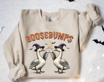 Może przedstawiać: Beżowa bluza z napisem "GOOSEBUMPS" w pomarańczowych literach. Grafika przedstawia dwie gęsi w czapce czarownicy, uderzające się pięściami. Nietoperze i półksiężyc również znajdują się w projekcie. Zabawna bluza z motywem Halloween.
