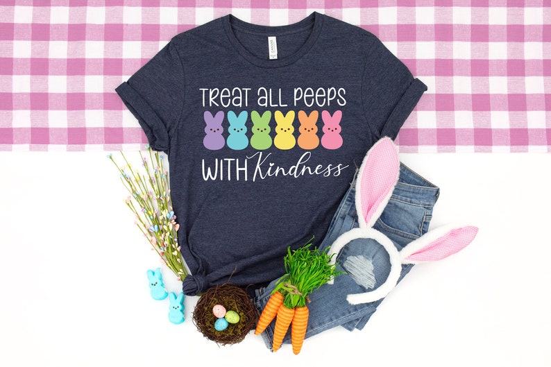 Może przedstawiać: Koszulka w kolorze granatowym z napisem "Treat all peeps with kindness" i rzędem tęczowych pianek marshmallow Peeps. Koszulka leży na r&oacute;żowo-białym obrusie w kratkę z parą uszu kr&oacute;liczych, gniazdem z jajkami wielkanocnymi i wiązką zielonej trawy.