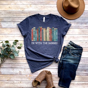 K&ouml;nnte beinhalten: Ein marineblaues T-Shirt mit einer Grafik eines B&uuml;cherregals voller verbotener B&uuml;cher. Der Text "I'm with the banned" ist unter der B&uuml;cherregalgrafik gedruckt.