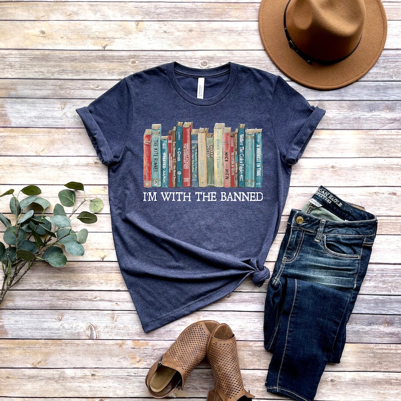 K&ouml;nnte beinhalten: Ein marineblaues T-Shirt mit einer Grafik eines B&uuml;cherregals voller verbotener B&uuml;cher. Der Text "I'm with the banned" ist unter der B&uuml;cherregalgrafik gedruckt.