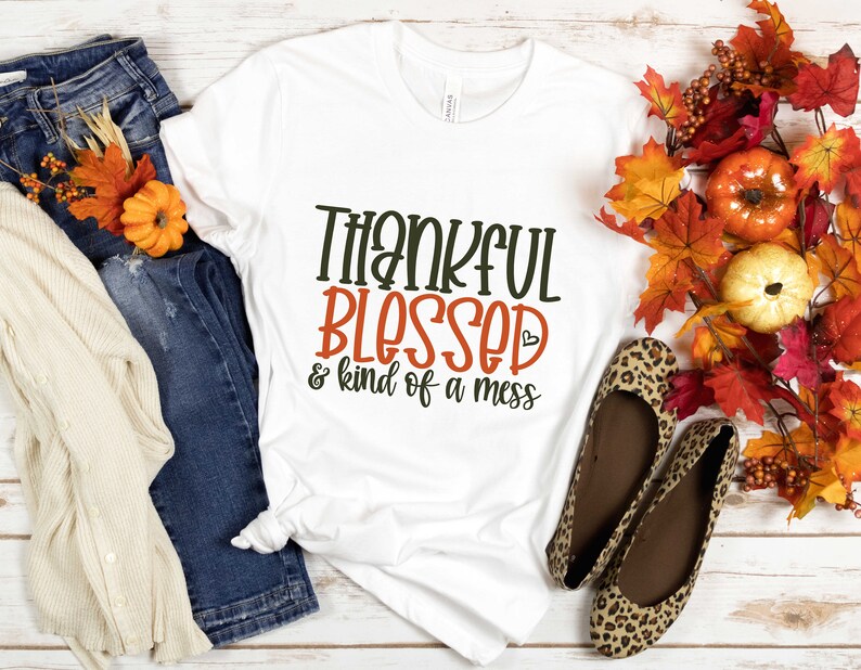 May include: White t-shirt with the text "Thankful Blessed & kind of a mess" in brown and orange lettering. The shirt is laying on a white wooden surface with a pair of leopard print flats, a pair of blue jeans, and a white cardigan.