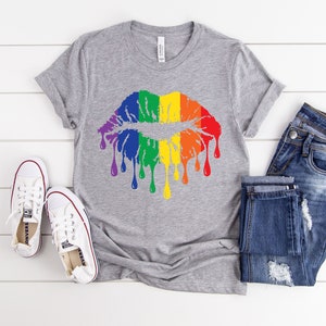 Op de afbeelding: Een grijs T-shirt met een regenboog-druppelende lippenontwerp. Het shirt wordt gecombineerd met een blauwe jeans en witte sneakers.
