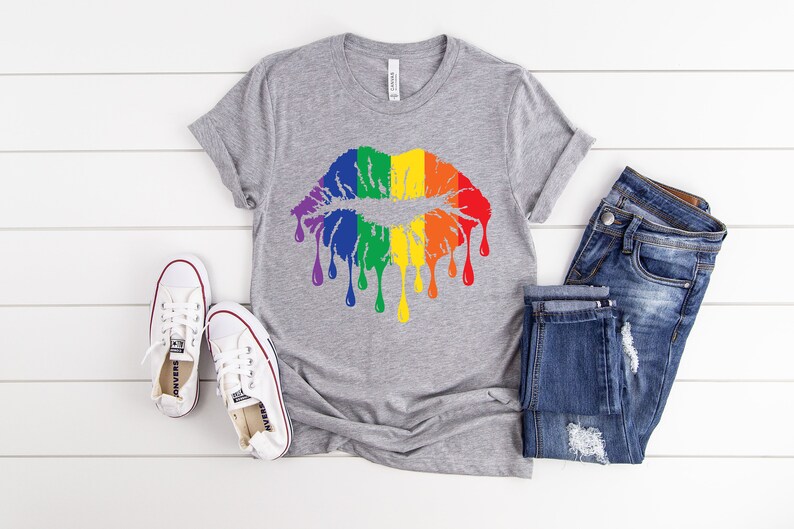 Op de afbeelding: Een grijs T-shirt met een regenboog-druppelende lippenontwerp. Het shirt wordt gecombineerd met een blauwe jeans en witte sneakers.