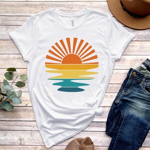May include: A white t-shirt with a retro sunset graphic featuring a sun with rays and a reflection in the water. The graphic is in shades of orange, yellow, blue, and green.